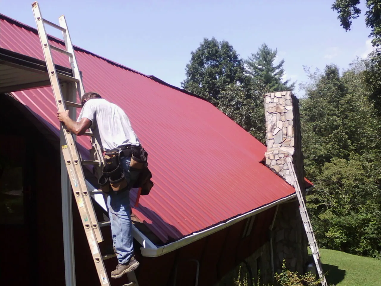 Roofer on a ladder beside a red metal roof and stone chimney — Soffit & Fascia Repair by Roofing Brothers in Gatlinburg
